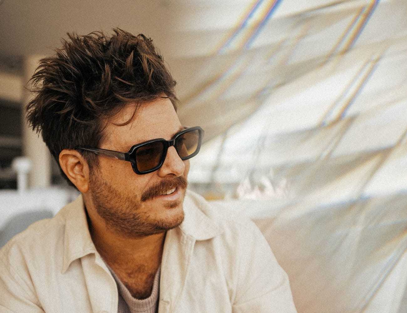 Man with sunglasses and a mustache, wearing a beige shirt, looks to the side in a sunny setting.