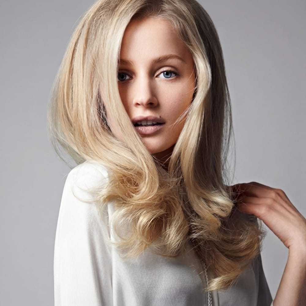 Woman with long blonde hair and white top, looking at camera against a gray background.