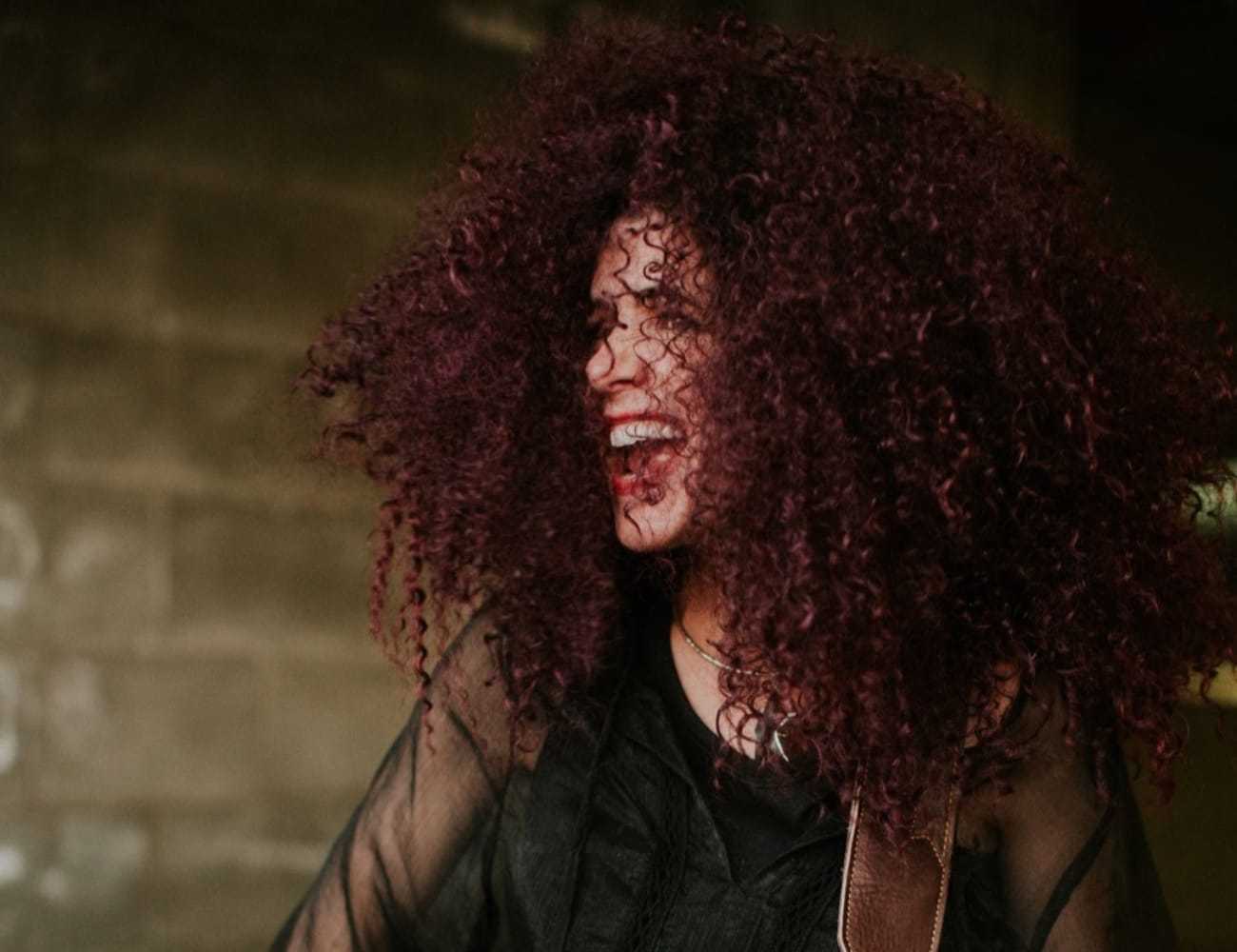 Singer energetically performing with curly hair flowing, against a dim brick backdrop.