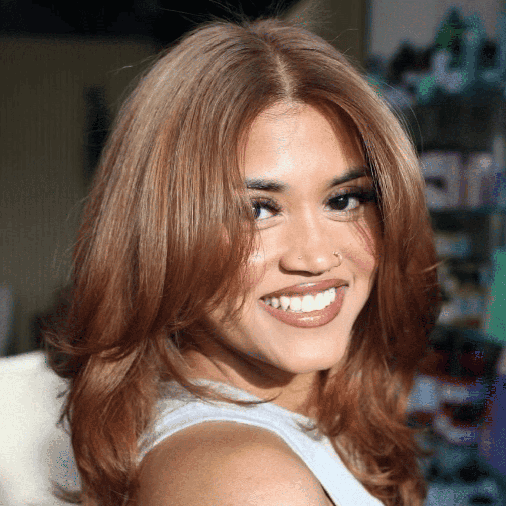 Smiling person with auburn hair and nose ring in a brightly lit room.