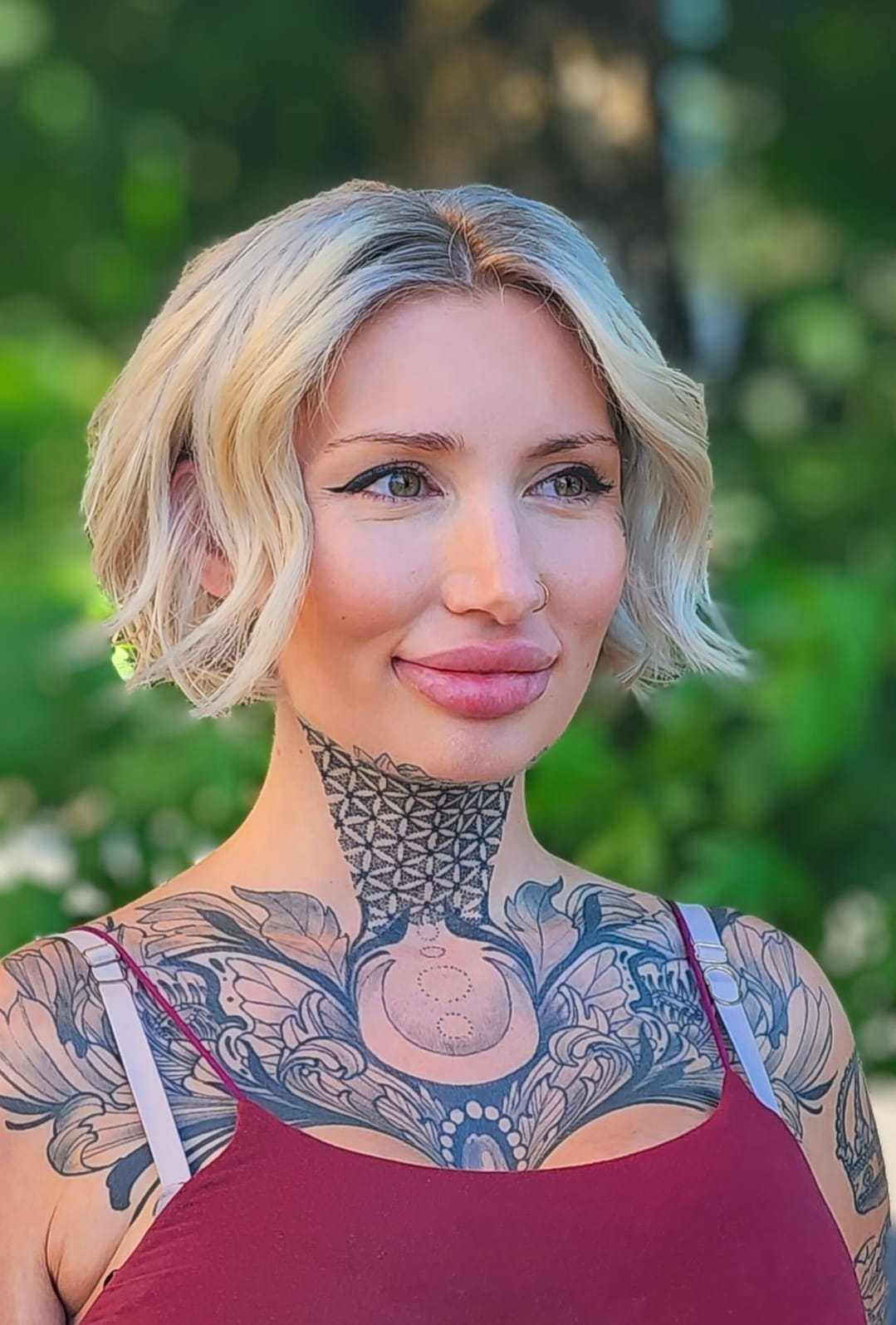 Tattooed woman with blonde bob haircut in outdoor setting.
