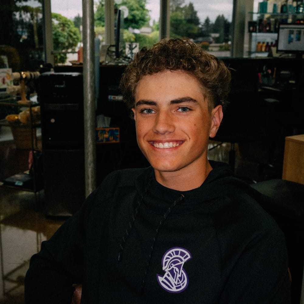 Young person smiling indoors, wearing a black hoodie with a logo.